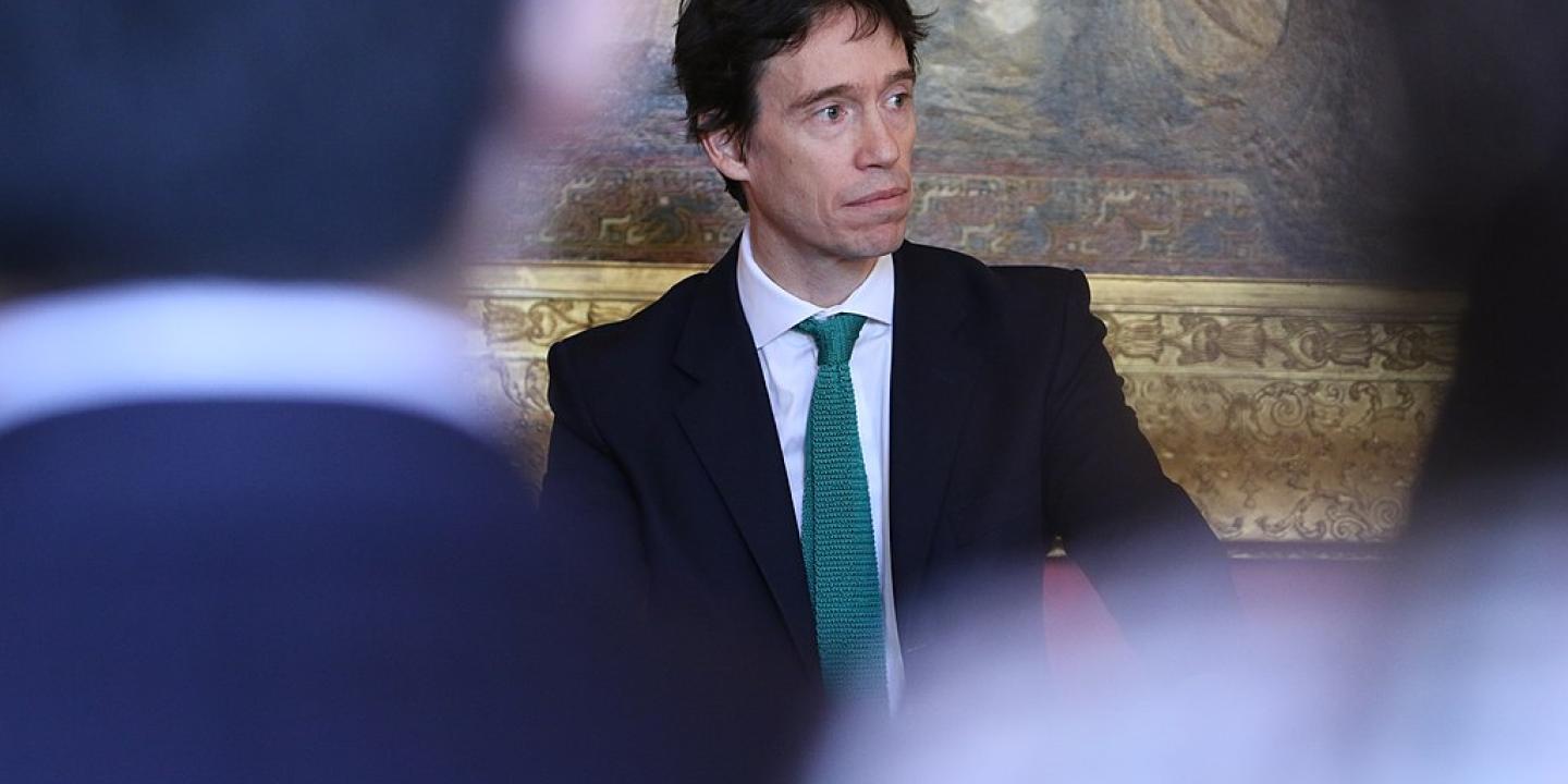 A suited politician stands looking pensive, framed by two out of focus audience members.