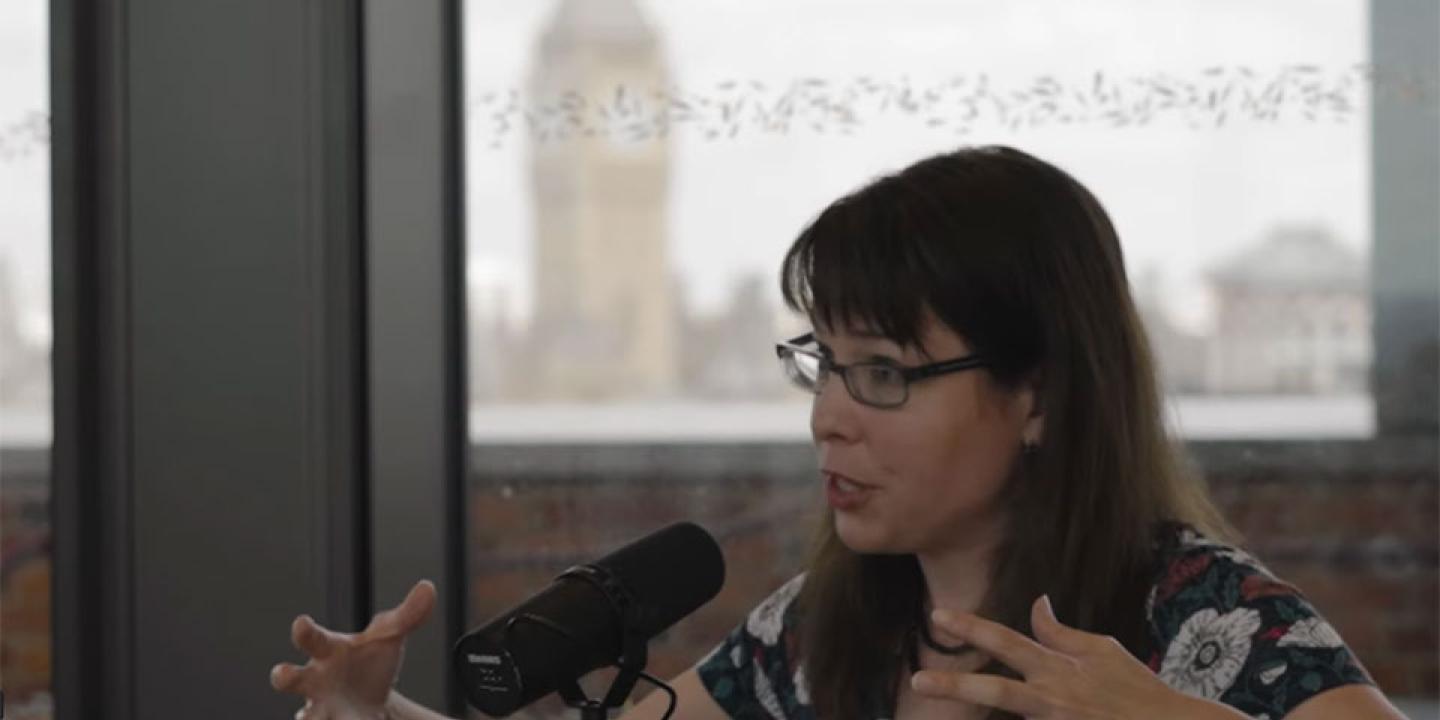 A woman seated at a table gestures with both hands while talking