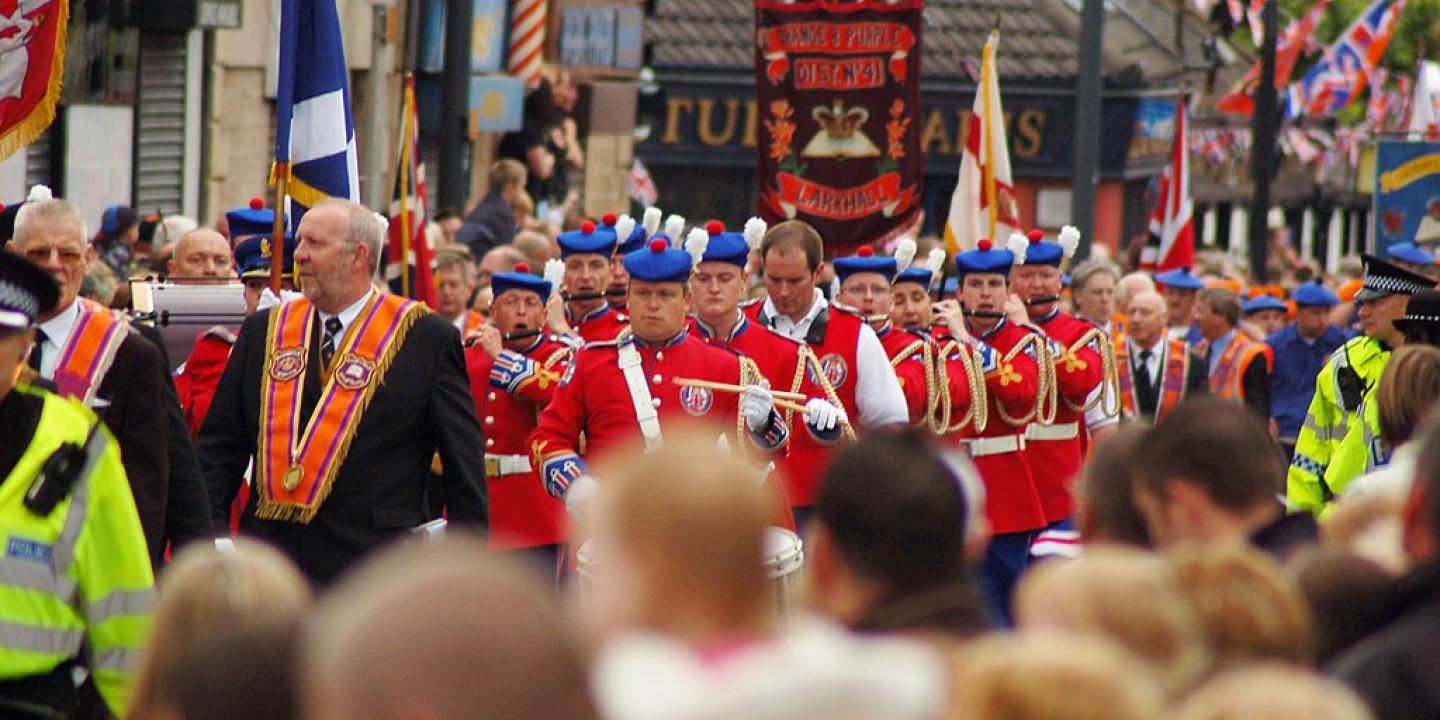 Across the heads of a roadside crowd, men wearing orange sashes and military band uniforms march along.