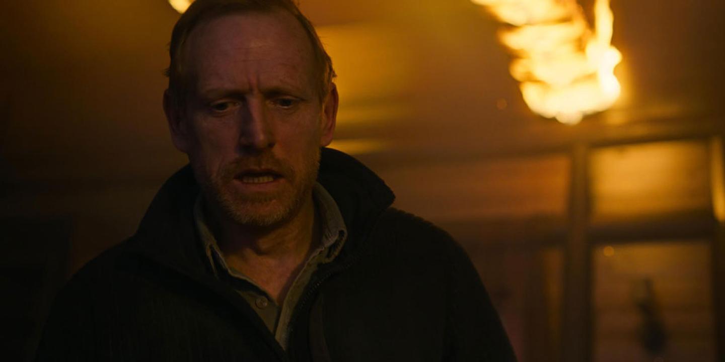 A crazed-looking man walks away from a burning backdrop.