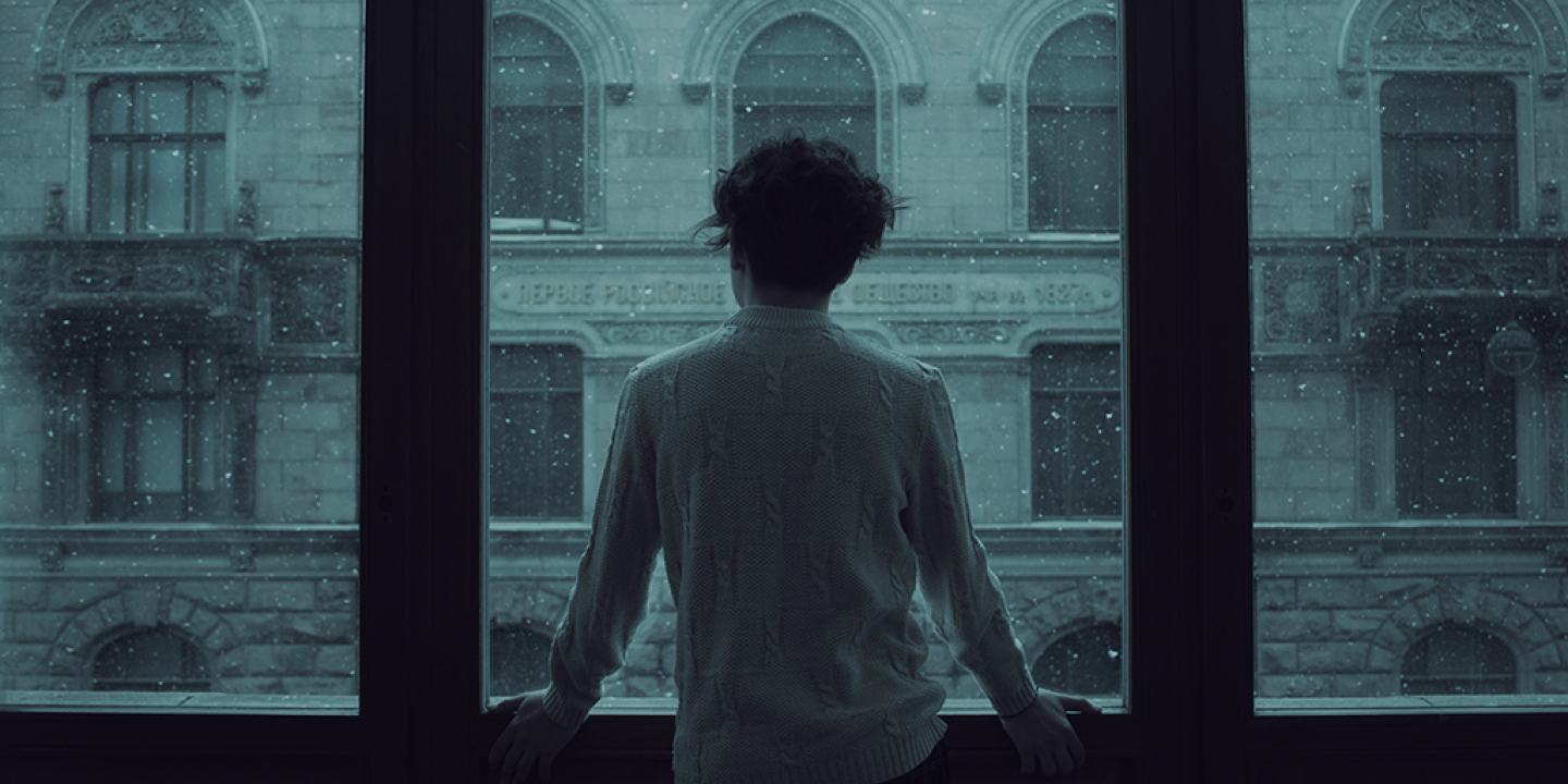 A person stand looking out a wide set of windows covered in rain.