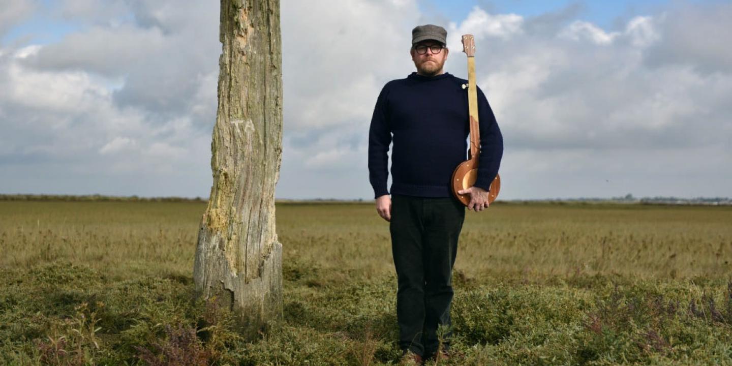 Standing on a salt marsh beside a wooden pillar, a man holds a banjo upright like a rifle.