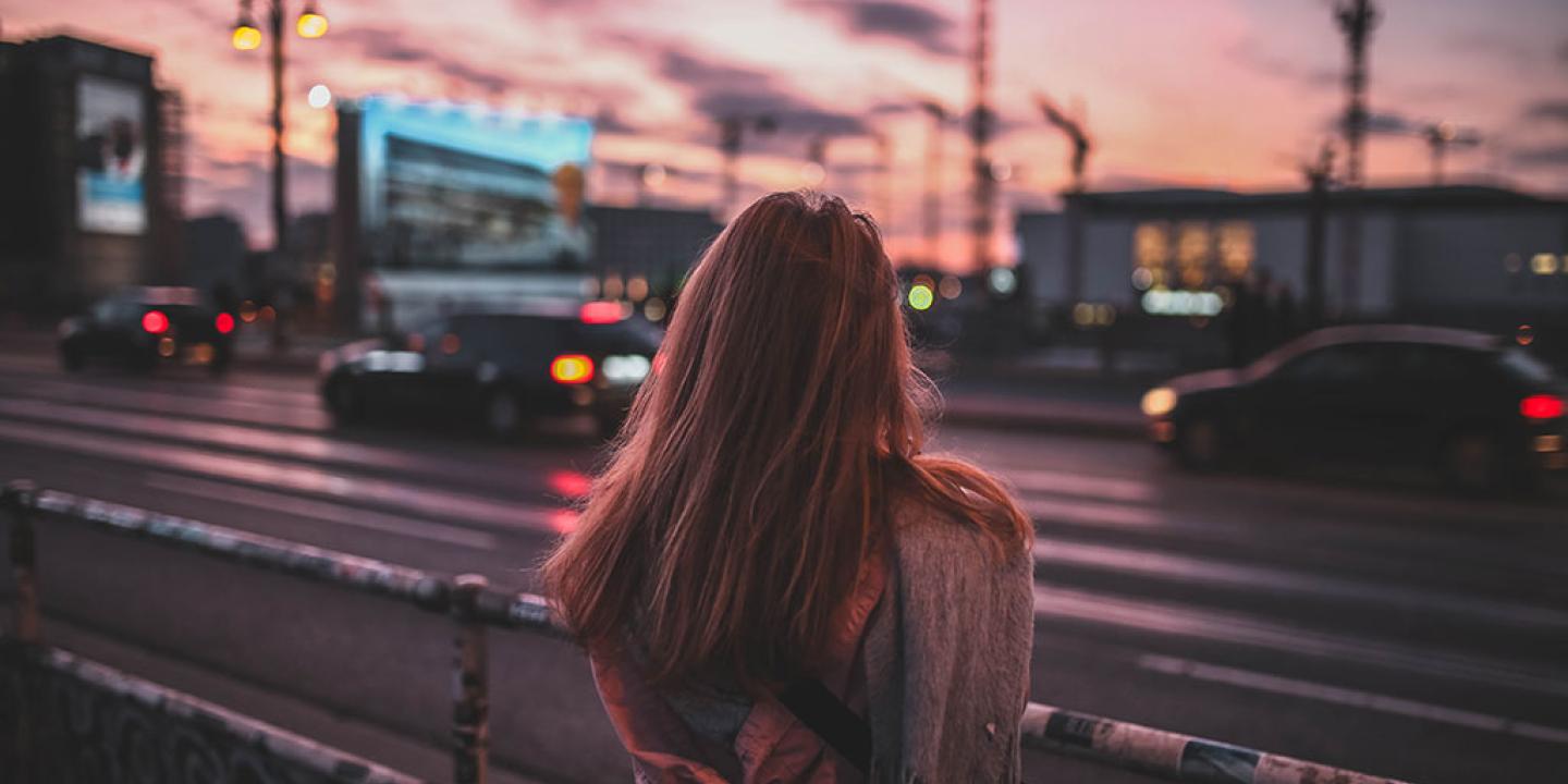 A woman stand beside a busy road with her back to the camera. There is a red sky behind her.