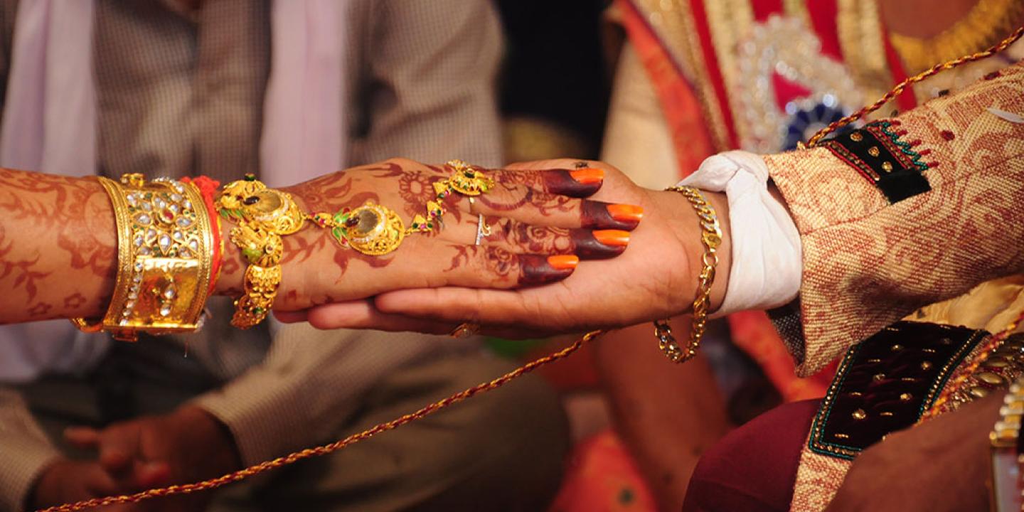 A close-up of a bride groom holding the brides hand. Her hand is henna tattooed and bears gold rings and bracelets. 