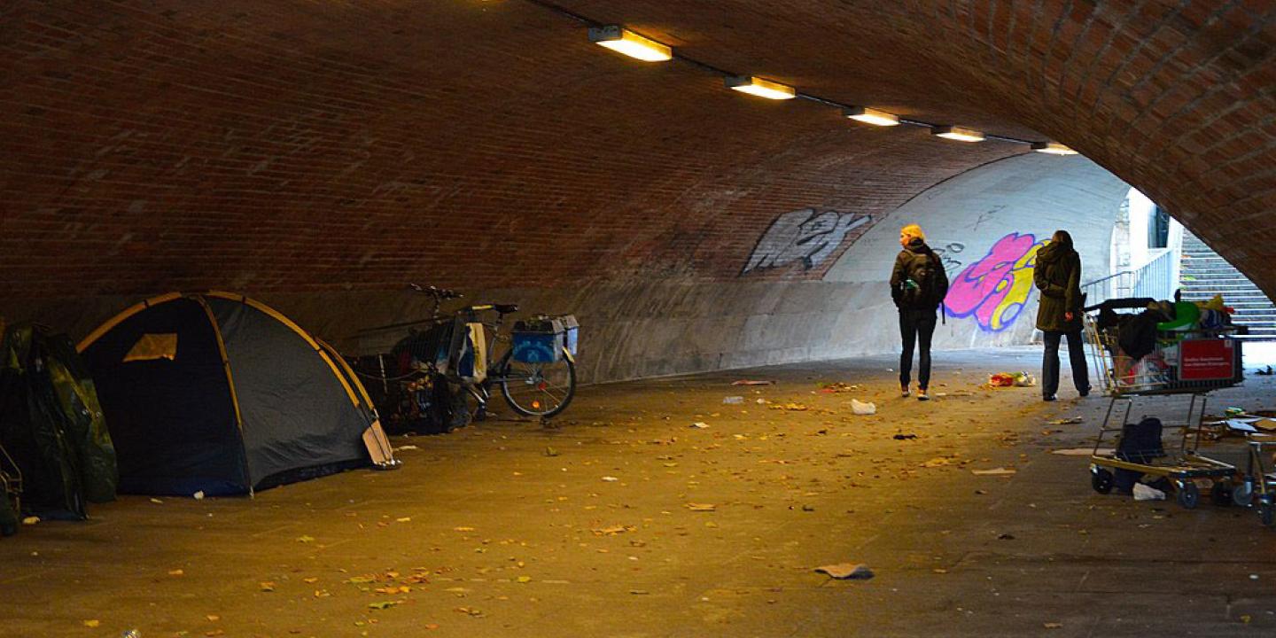 In an underpass a pedestrian passes and look at the tent of a homeless person.