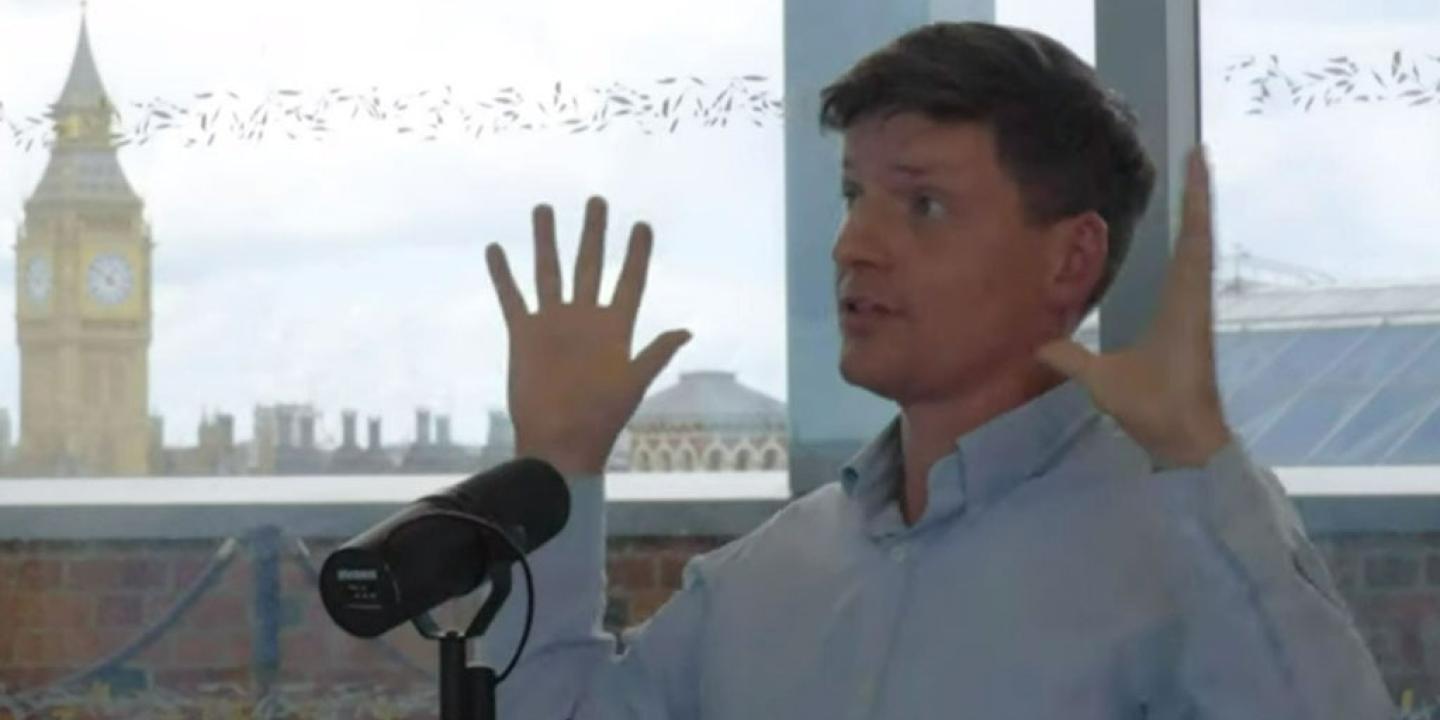 A man sits at a table recording a podcast and holds his hands up for emphasis.