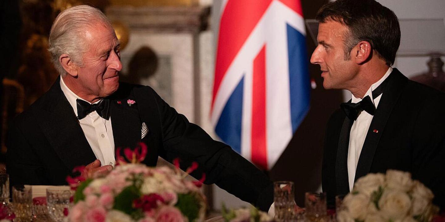 King Charles in a black dinner suit talks and gestures to President Macron who is similarly dressed.