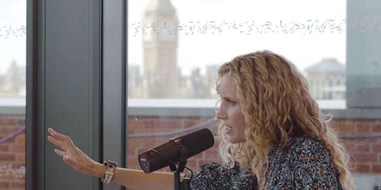 A seated podcast guest talks into a mic while stretching out a hand for emphasis.