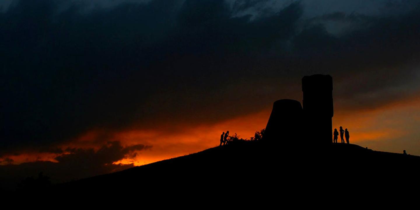 A sunset dramatically silhouette's a ruined tower and people at its base.