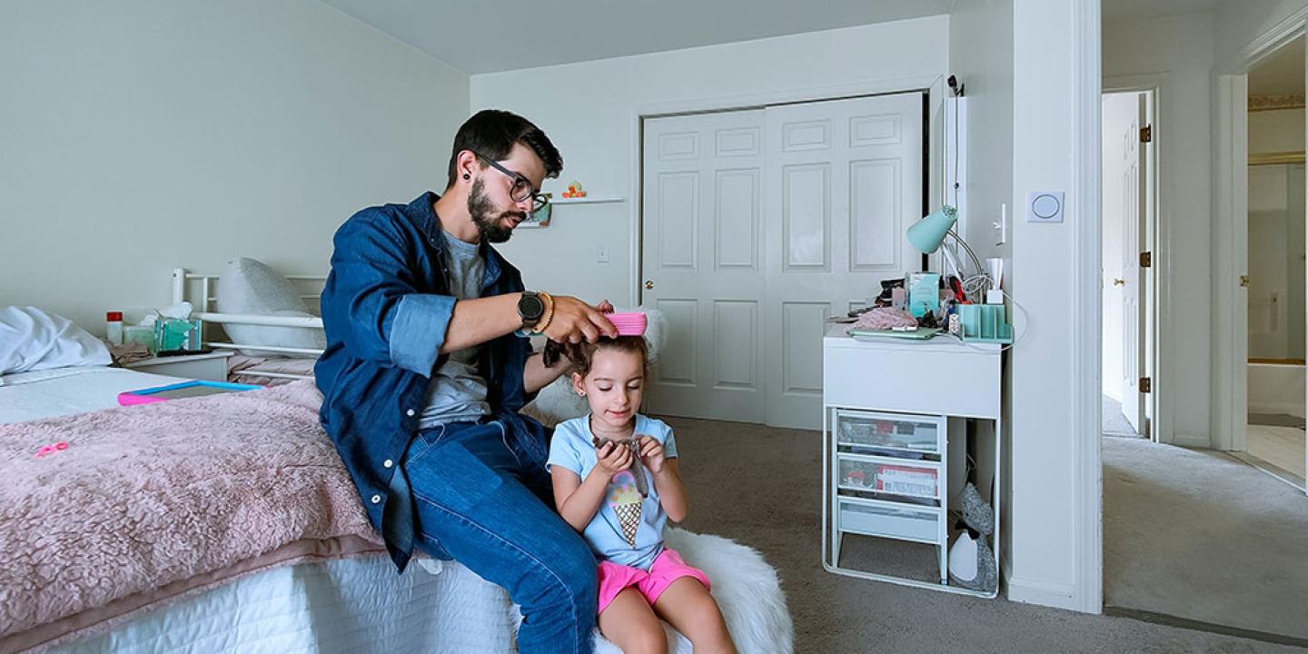 A father sits on a bed and fixes the hair of his daughter standing in front of him