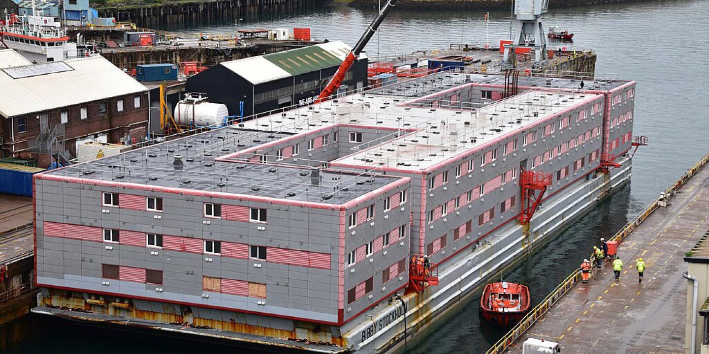 A grey multi-story accommodation barge floats beside a dock.
