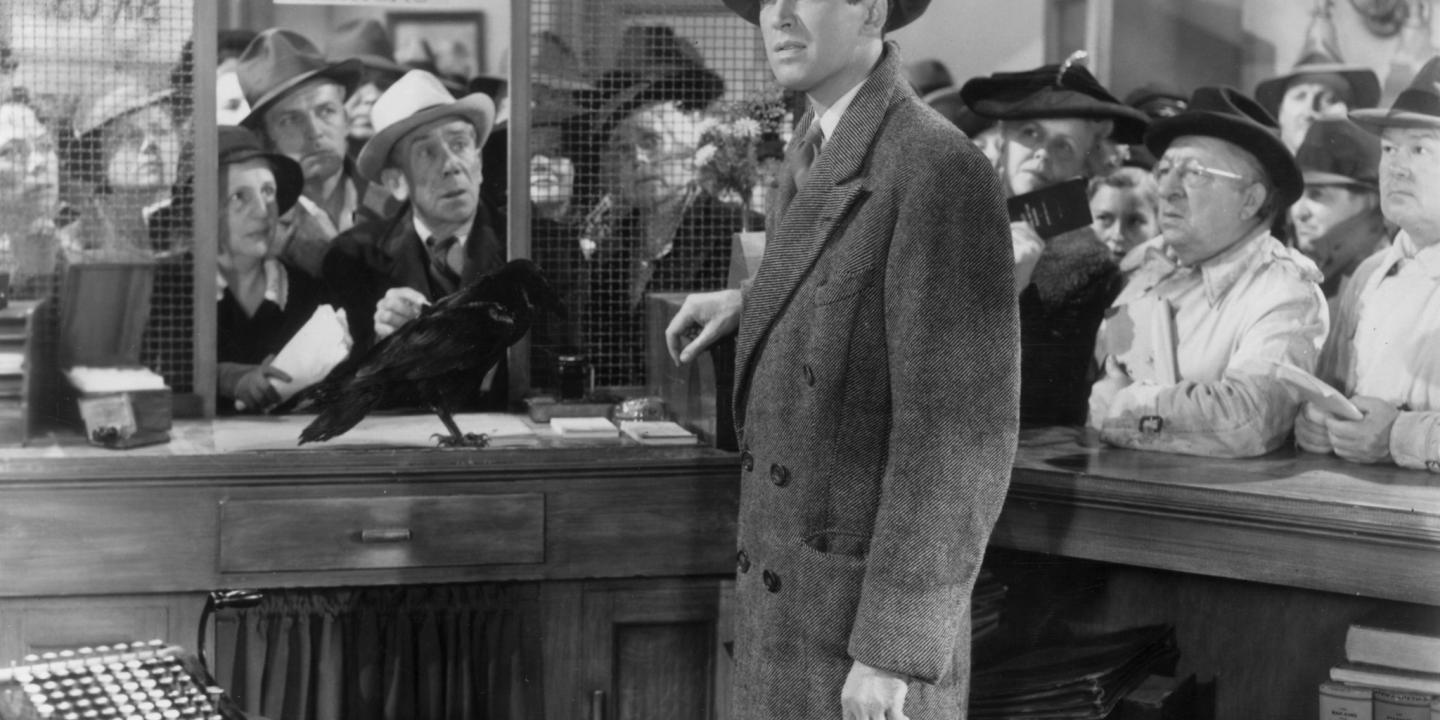 A man stands one side of a bank counter while others, on the other side, look hopefully at him.