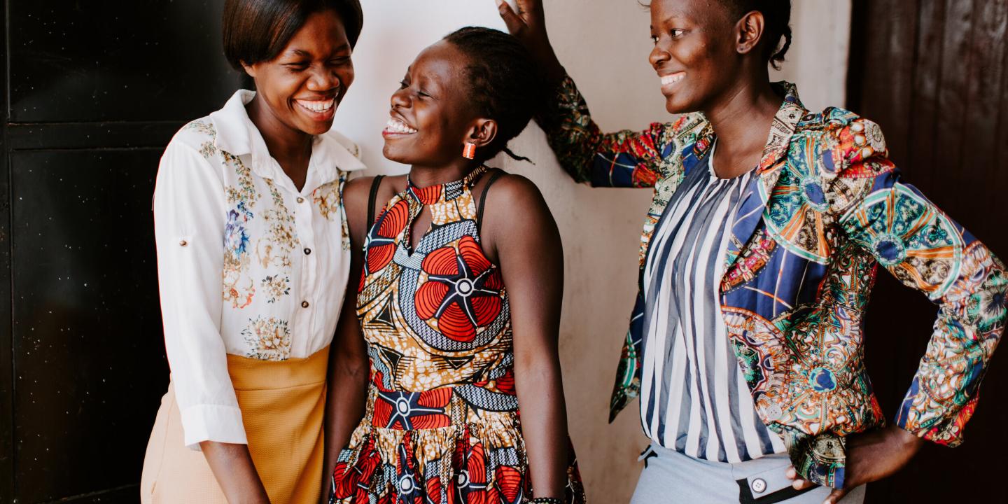 Three women stand, two lean into each other sharing a joke, while the other laughs too.