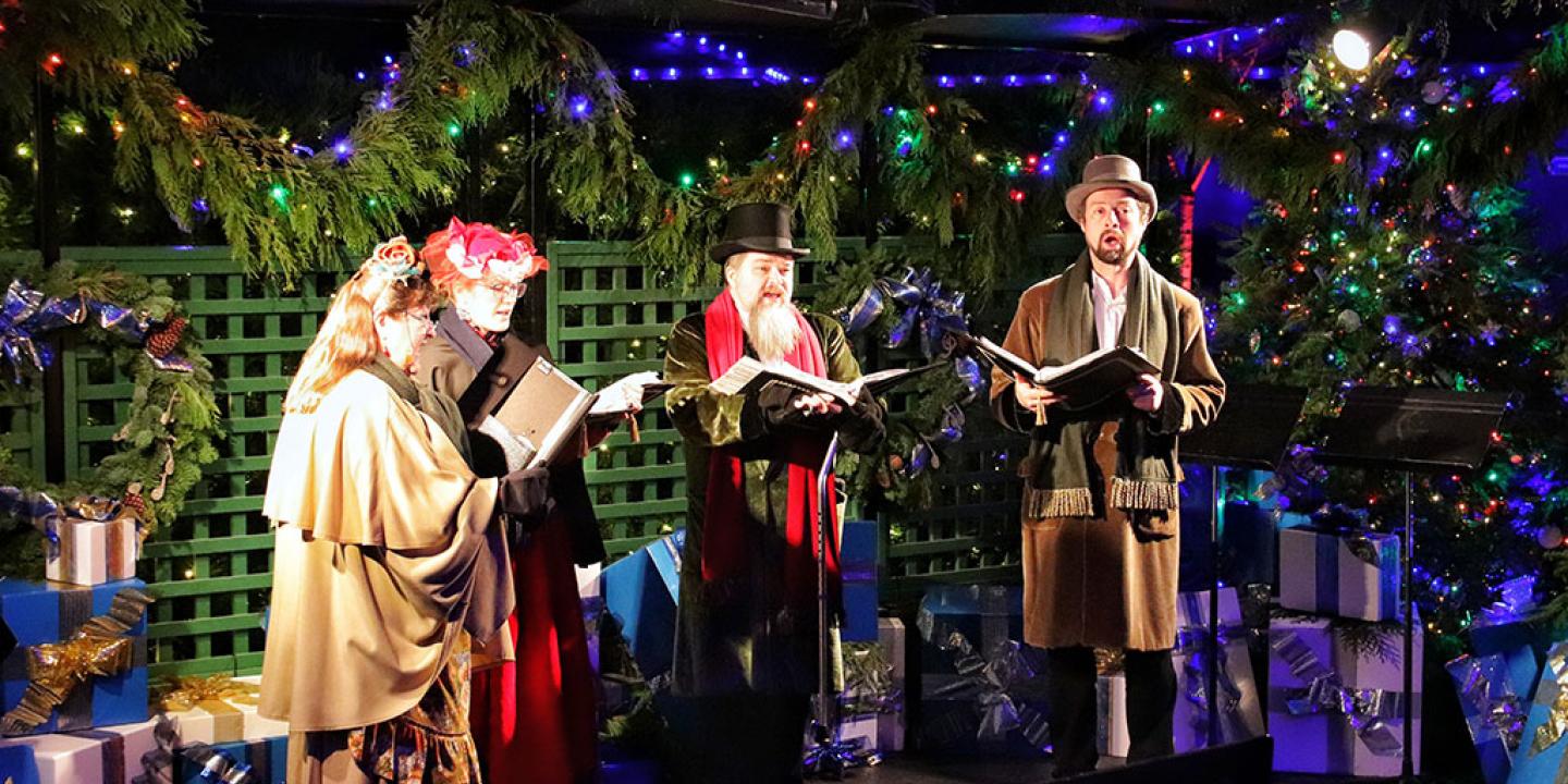 Dressed in Victorian clothes, a group of carol singers stand and sing amid Christmas foliage.