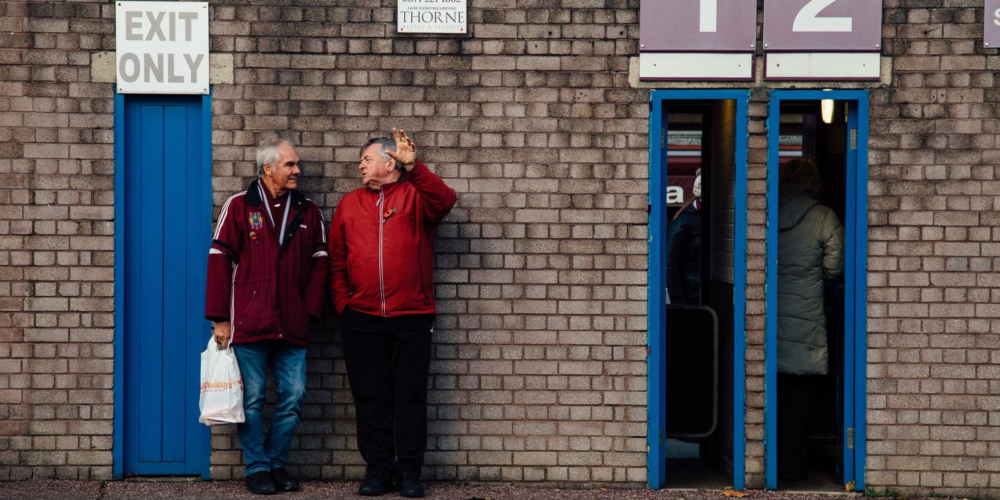 Two older men lean agains the wall of a football club's entrance, next to thin open doorways.