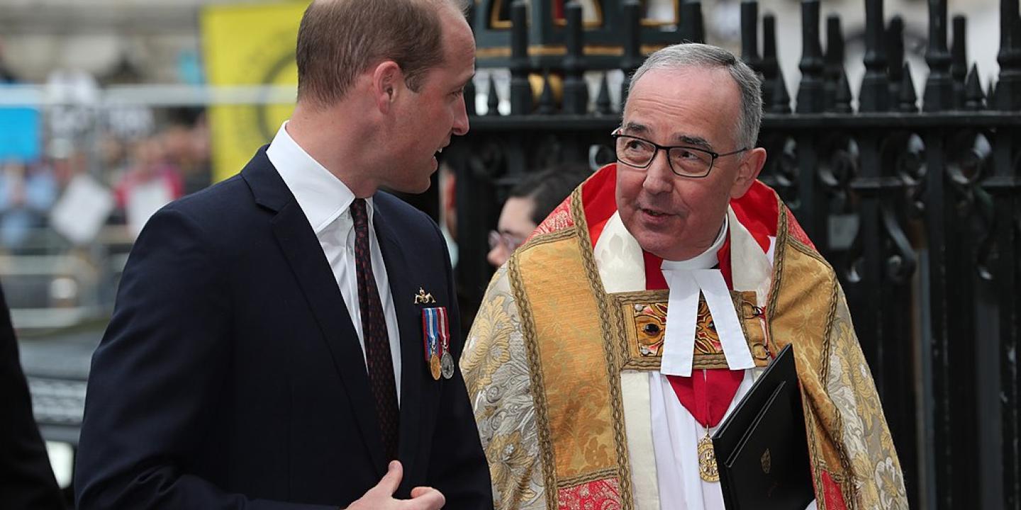 A young man wearing a dark suit talks to a minister wearing regalia.