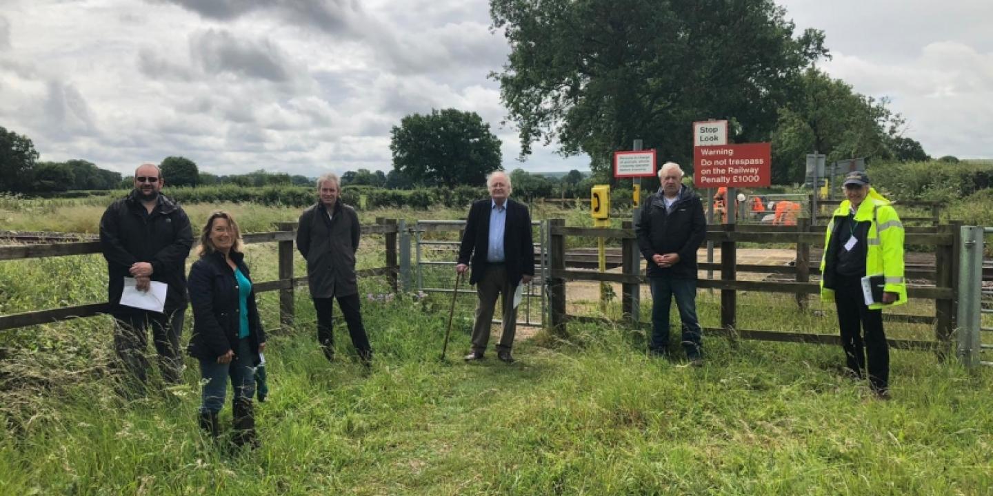 A group of people stand in a field by a fence and a railway footcrossing.