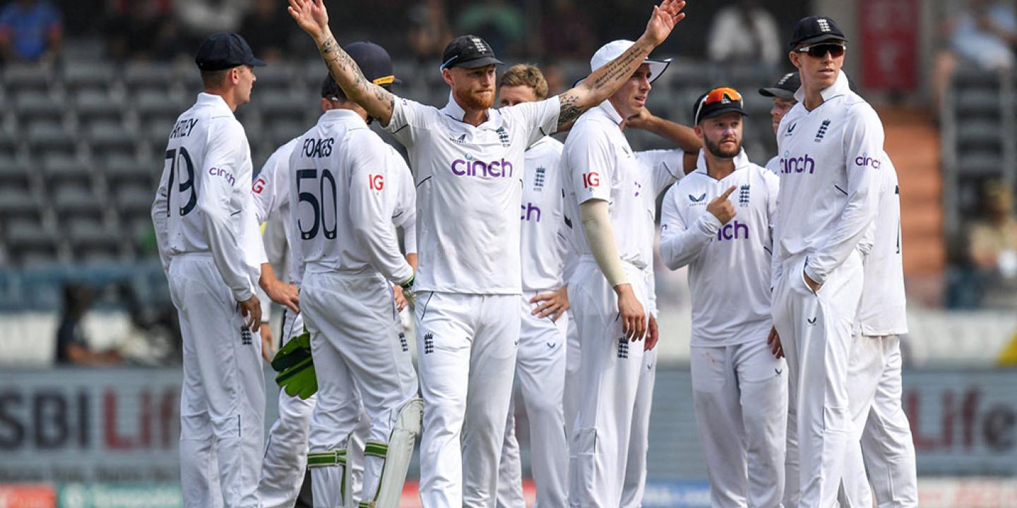 A gaggle of cricket players, dressed in whites, stand on the field. One raises there arm