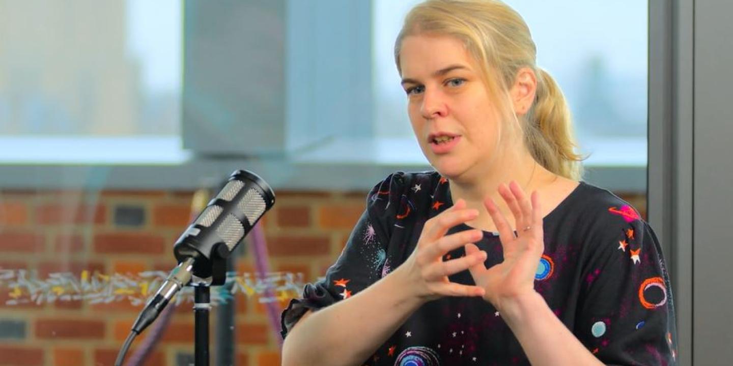 a woman sits at a table, with a mic in front of her, talking and holding her hands together in front of her.