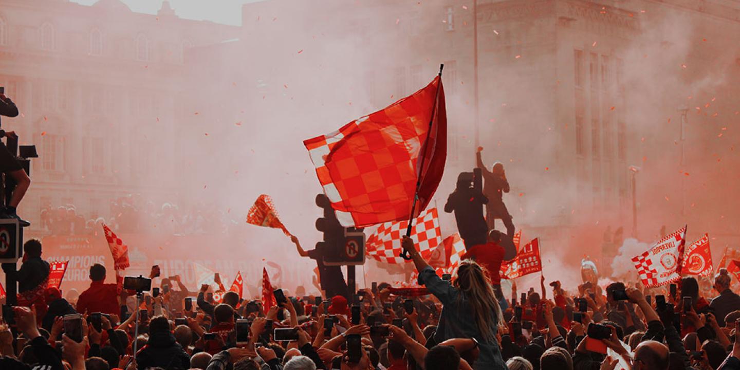 Silouhetted by red flare smoke, celebrating footballs wave red flags.