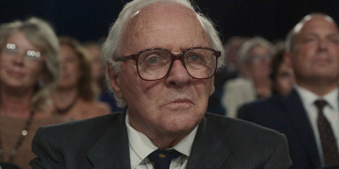 An old man wearing a suit and tie sits in a TV audience as people stand around him.