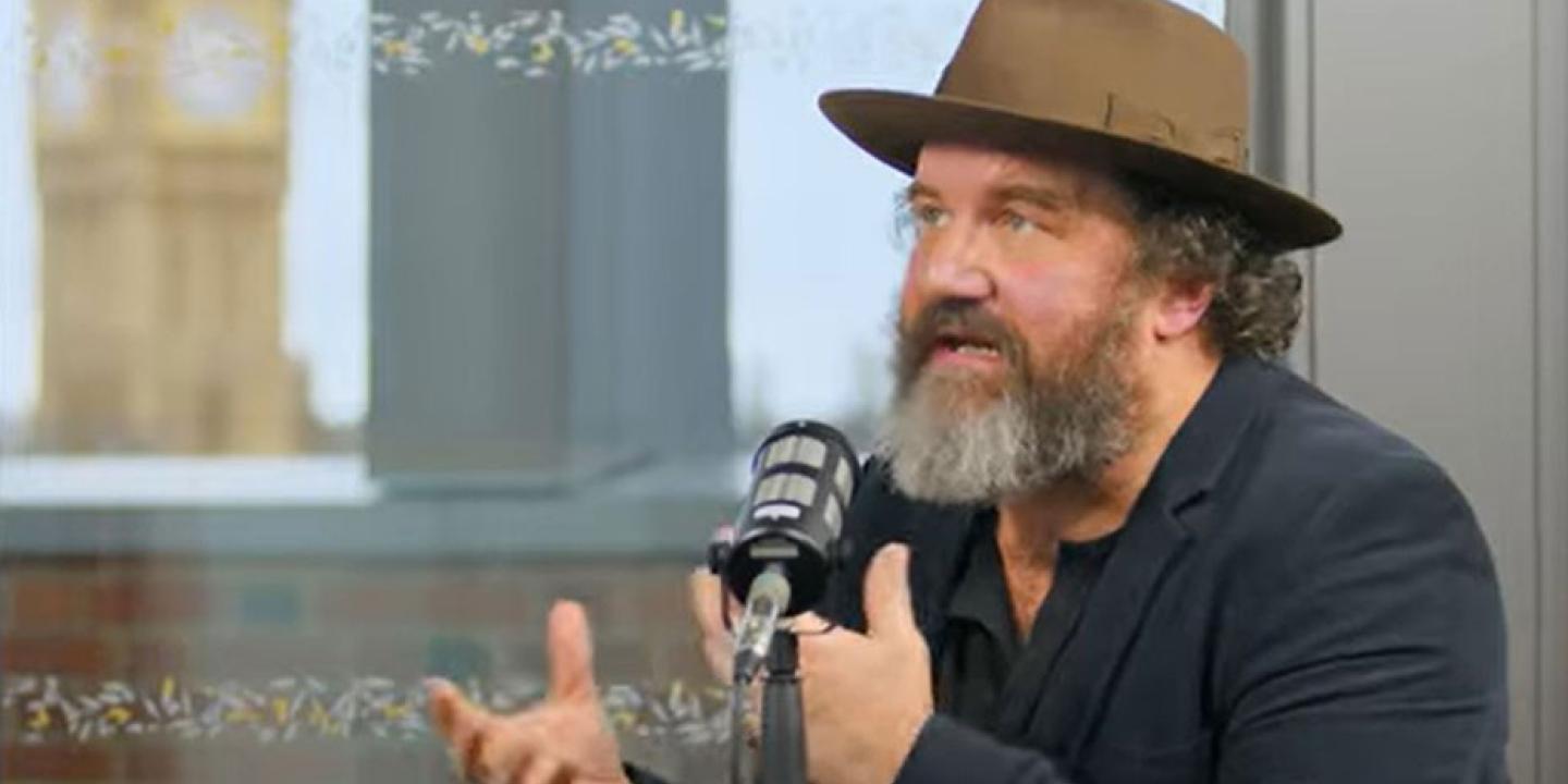 A man wearing a hat sits at a table talking and raises both hands in front of himself to gesture