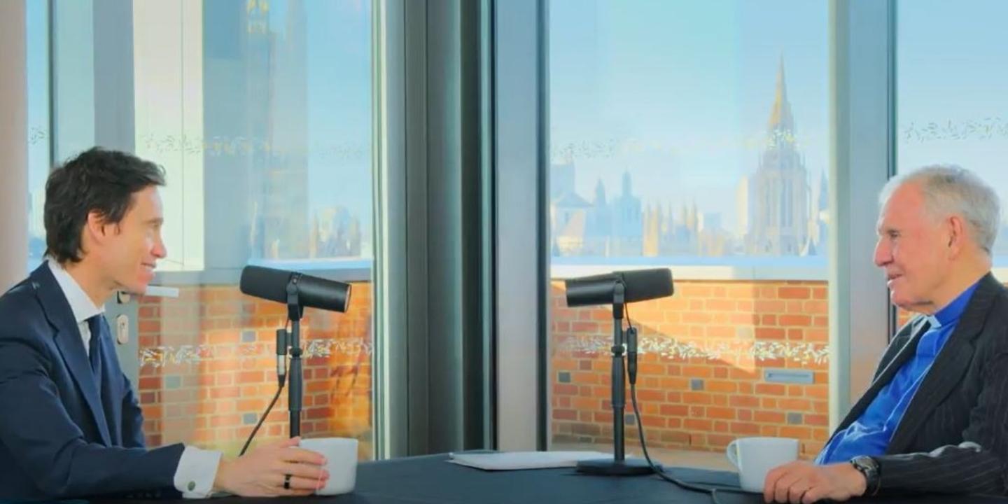 two people sit opposite and converse, microphones are on the table.