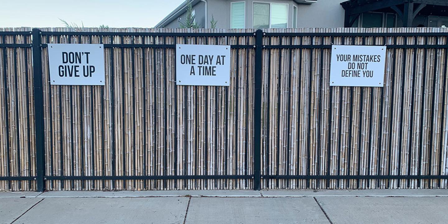 Three signs attached to a fence read: Don't Give Up, One Day At A Time, and Your Mistakes Don't Define You.