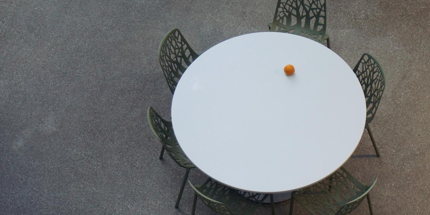 a round table with empty chairs is seen from above. An orange is the only item on it.