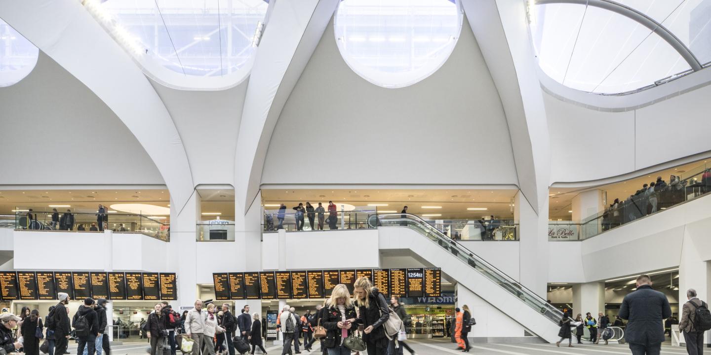 A airy and light station concourse in which people stand and look at a long set of travel information screen.
