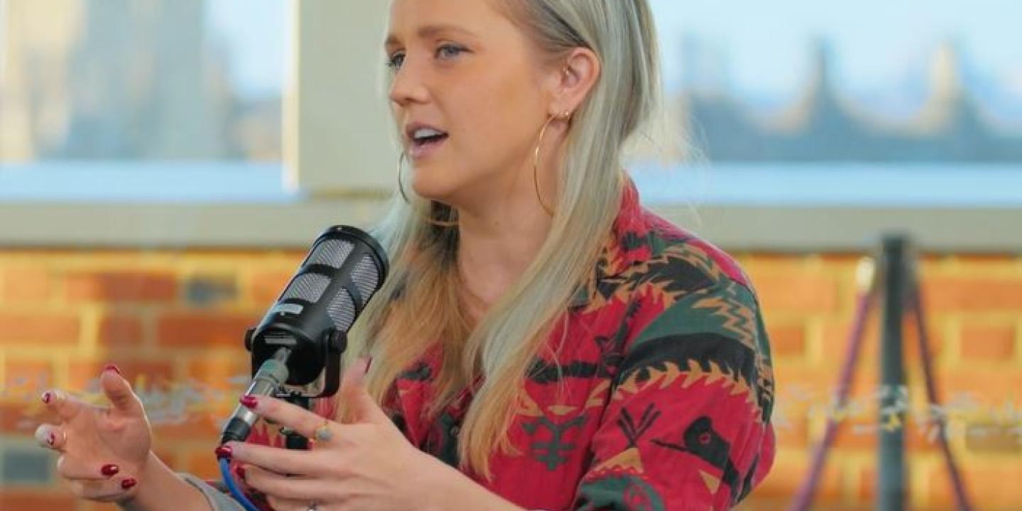 A sitting woman leans forward with raised open hands while at a table with a microphone on it.