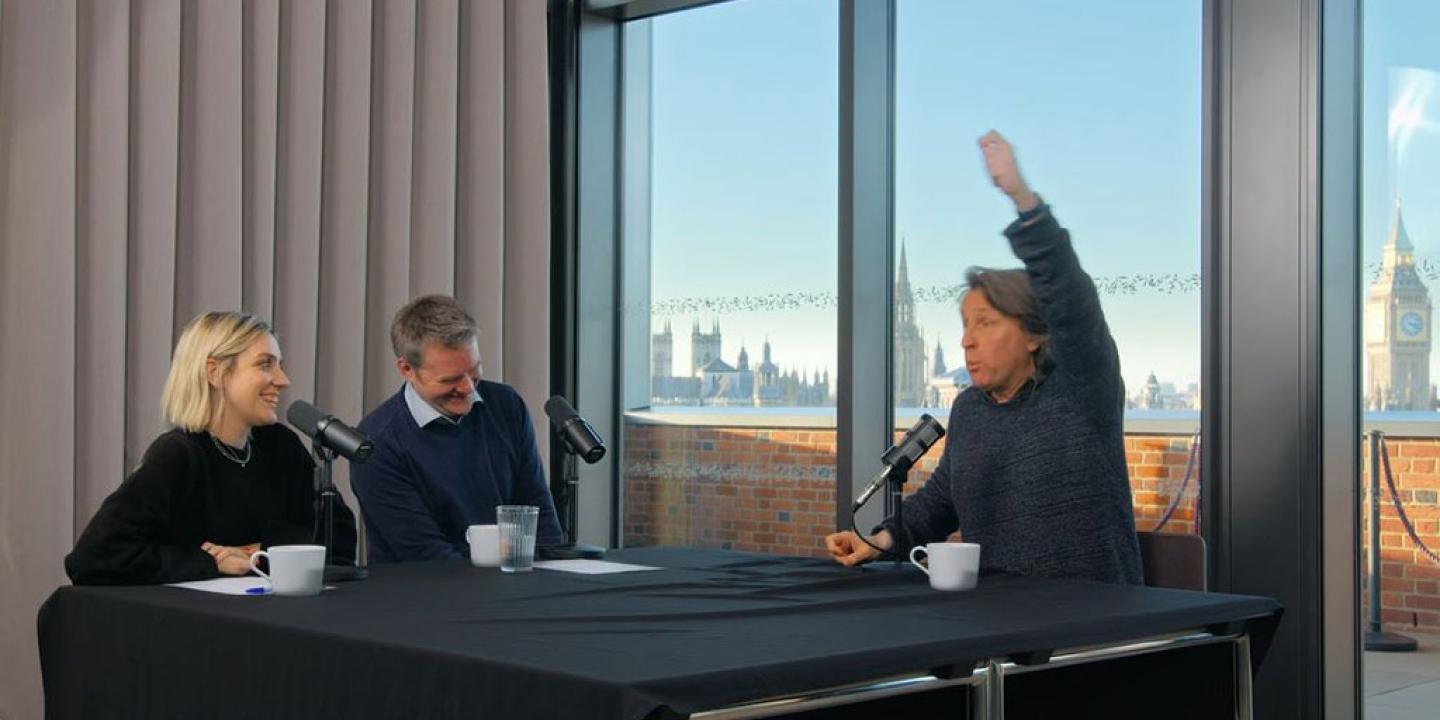 A sitting man raises one hand above his head to emphasise a point, while two other people looking on are laughing.