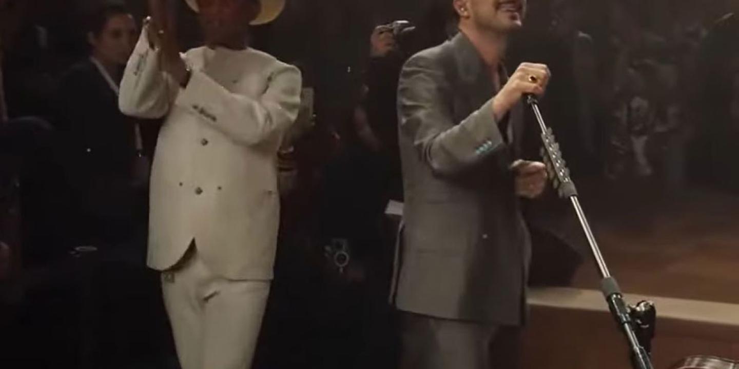 Two singers peform together. One in a white suit and stetson claps their hands. The other tilts the mic stand.