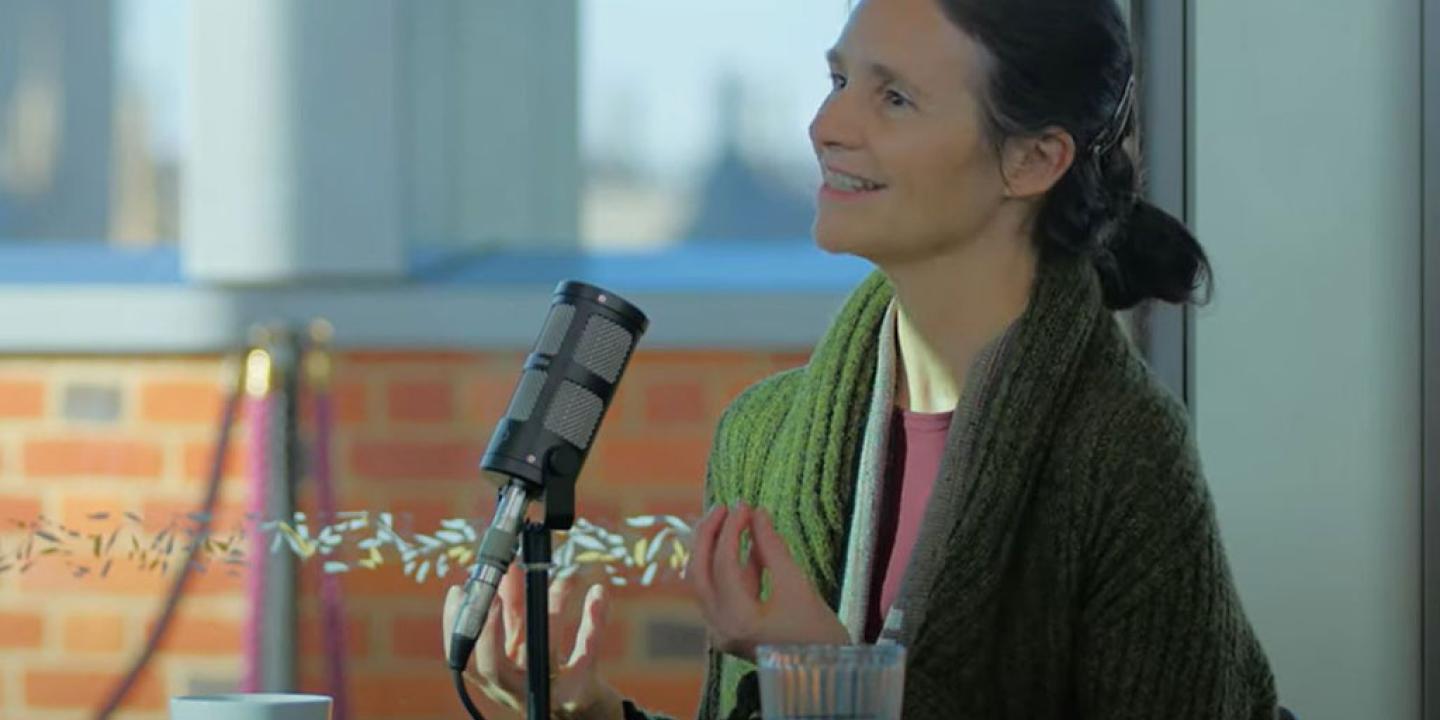 a woman sits at a table, with a mic in front of her, talking and holding her hands out in front of her.