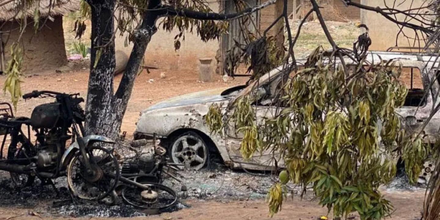 A burnt out motor cycle and car stand amid charred debris in a dusty compound.