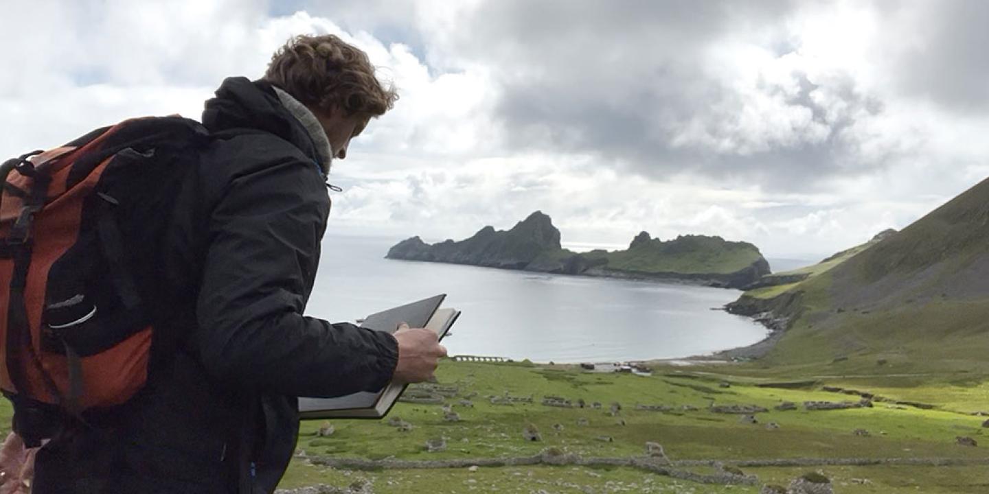 An artist holds a sketchbook while standing overlooking a deserted village by a bay, sided by jagged cliffs.