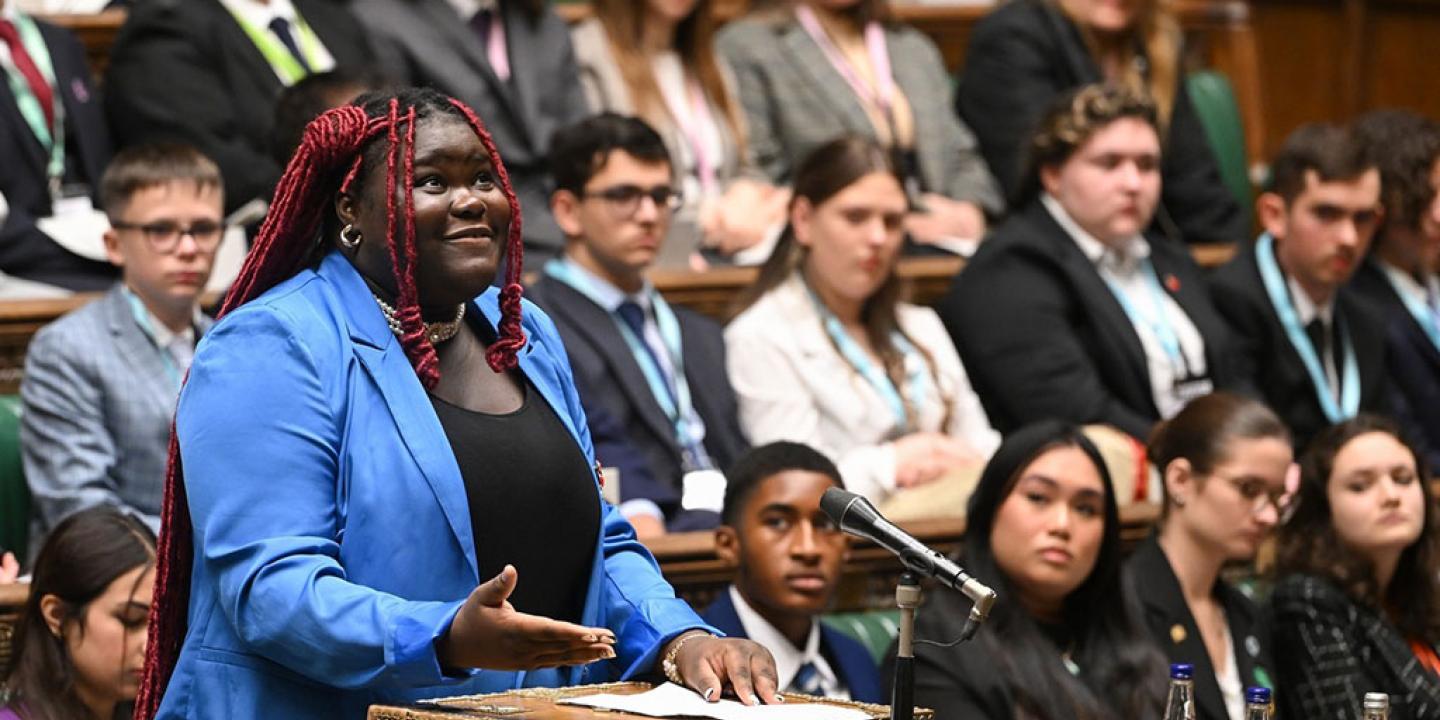 A young woman in a blue suit stands at a wooden box in a parliamentary debating chamber looking upward while speaking. 