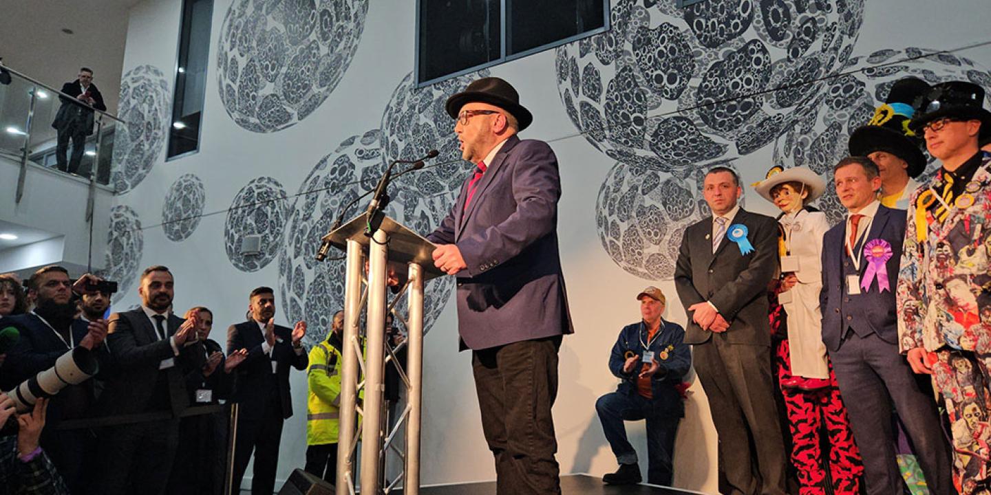 A winning candidate at an election address the audience from a lectern while the loosing candidates look on