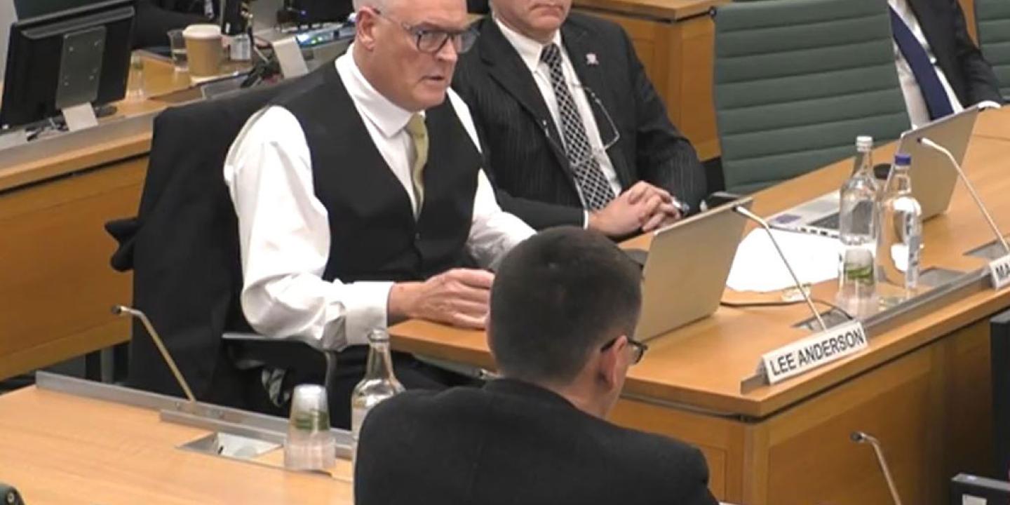 A man wearing a waist coats sits at a desk and ask a questions of a panel of people with their backs facing us.