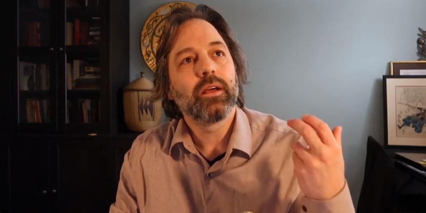 A man sits at a desk and looks up and gestures with his hand.