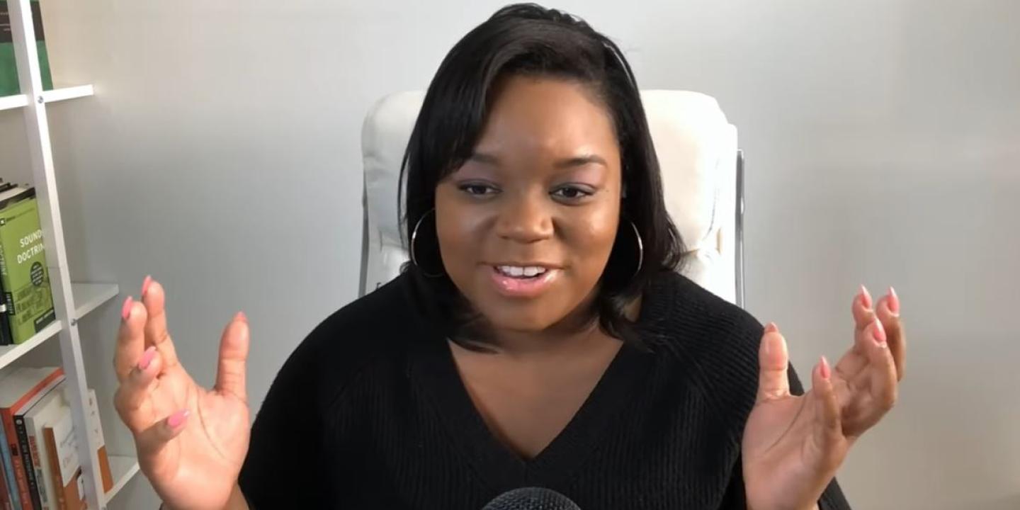 A woman sits and speaks into a desktop microphone and holds both hands up and out expressively.