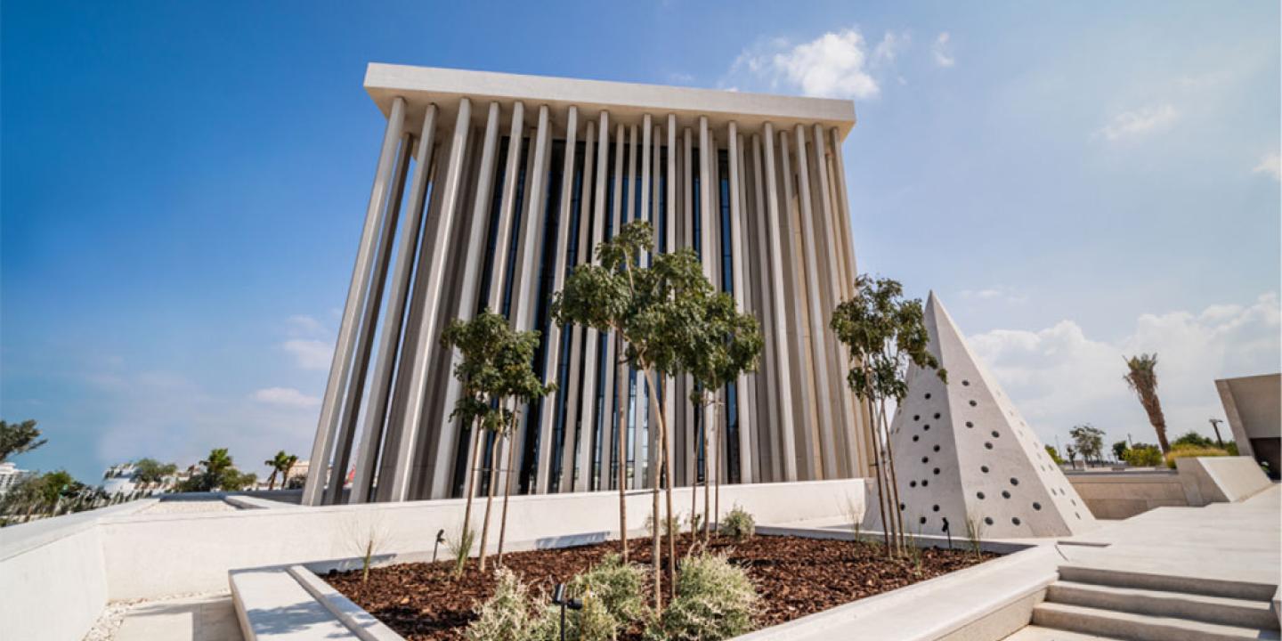 A large, modern cubiod building with a series of thin external pillars is seen from low down against a sunny blue sky 