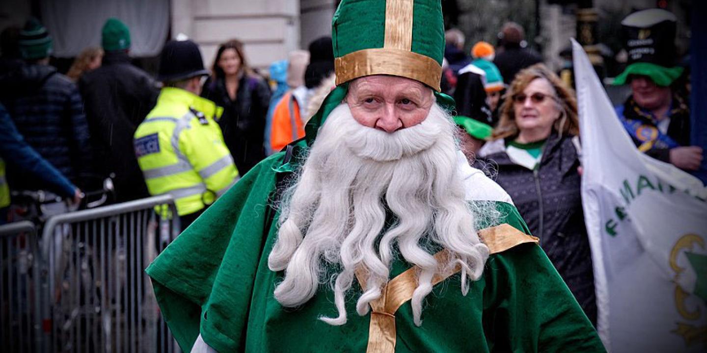 A parade particpant dressed as a bishop in green vestments with a false beard walks down a street.