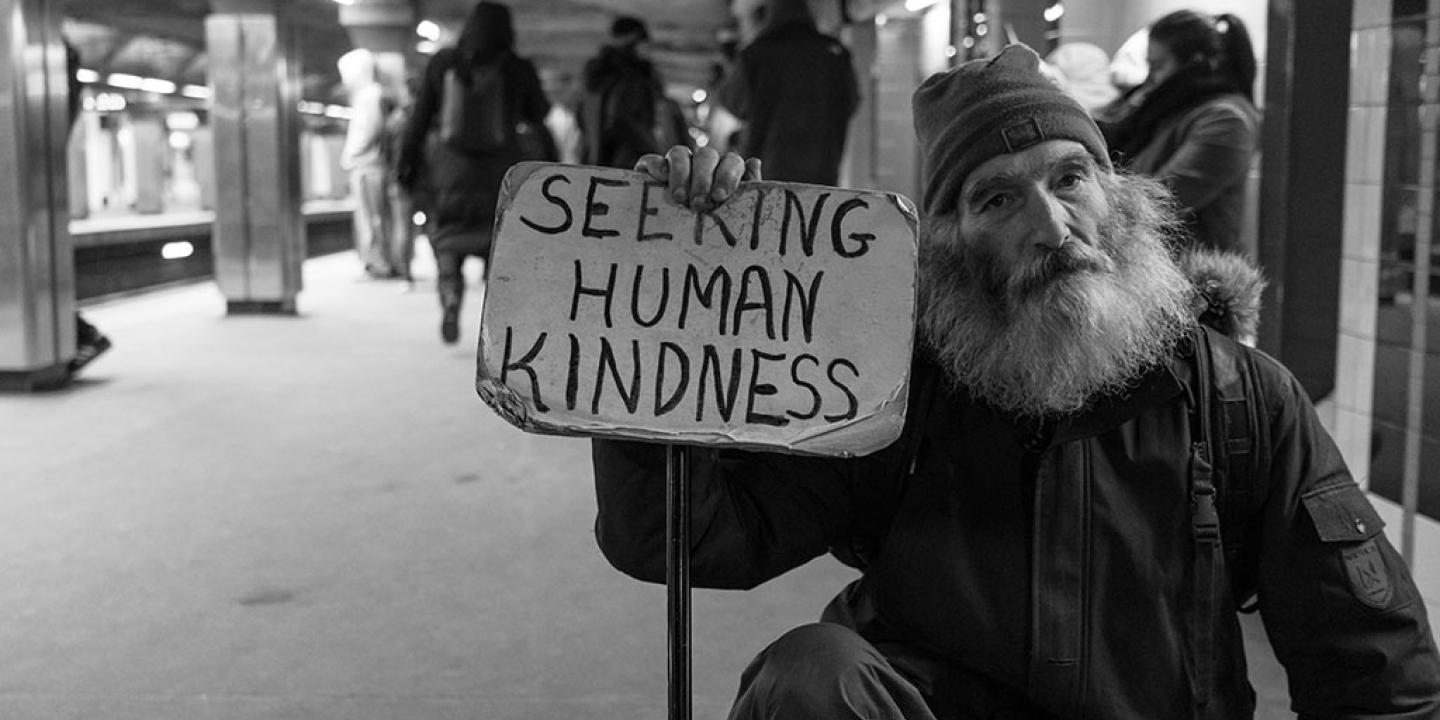 a man in a wheelchair sits in a subway station holding a sign reading 'seeking human kindness'.