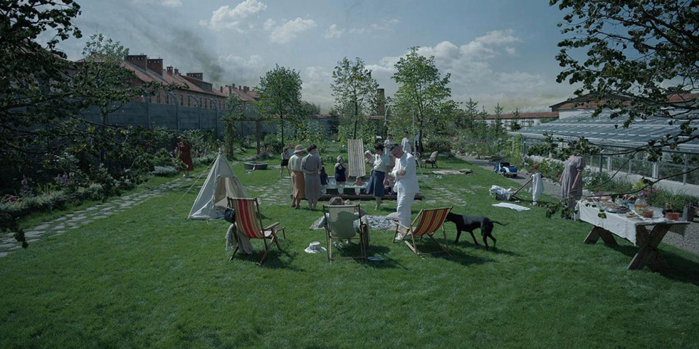 in an immaculate garden a family play in and around a small swimming pool. Beyond the garden wall, a barracks is visble with crematorium smoke rising beyond it.