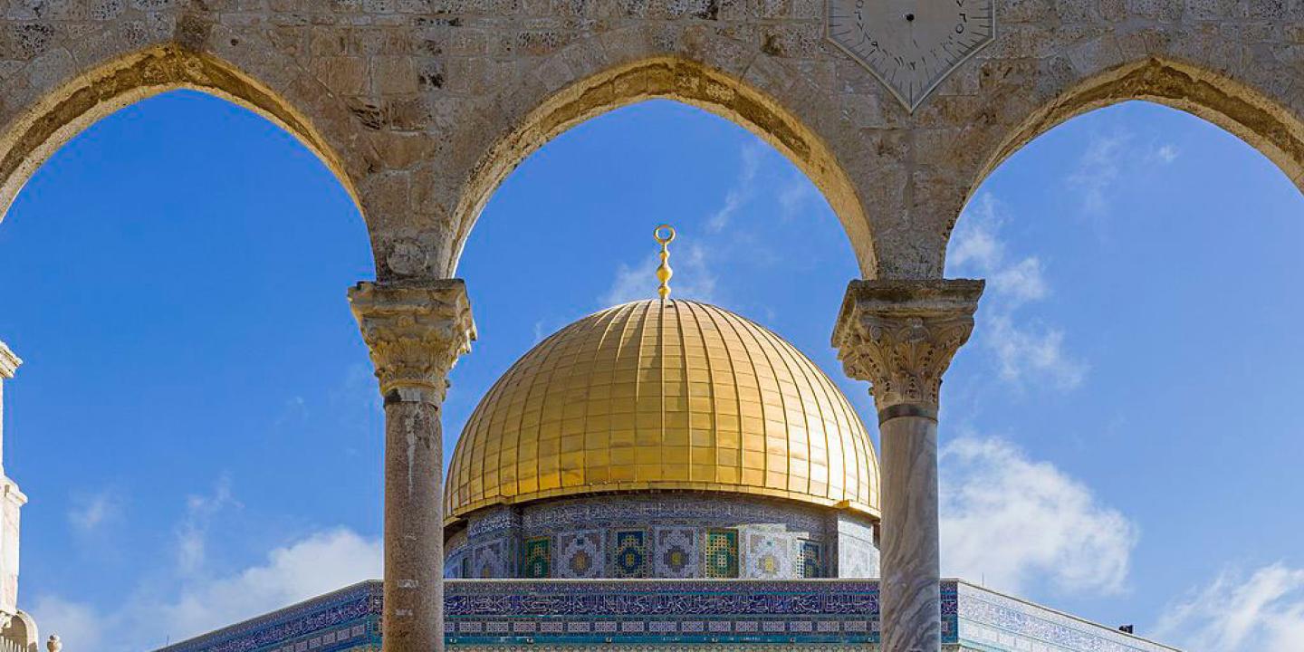 A gold-domed, blue-walled octagonal mosque seen through a row of arches.