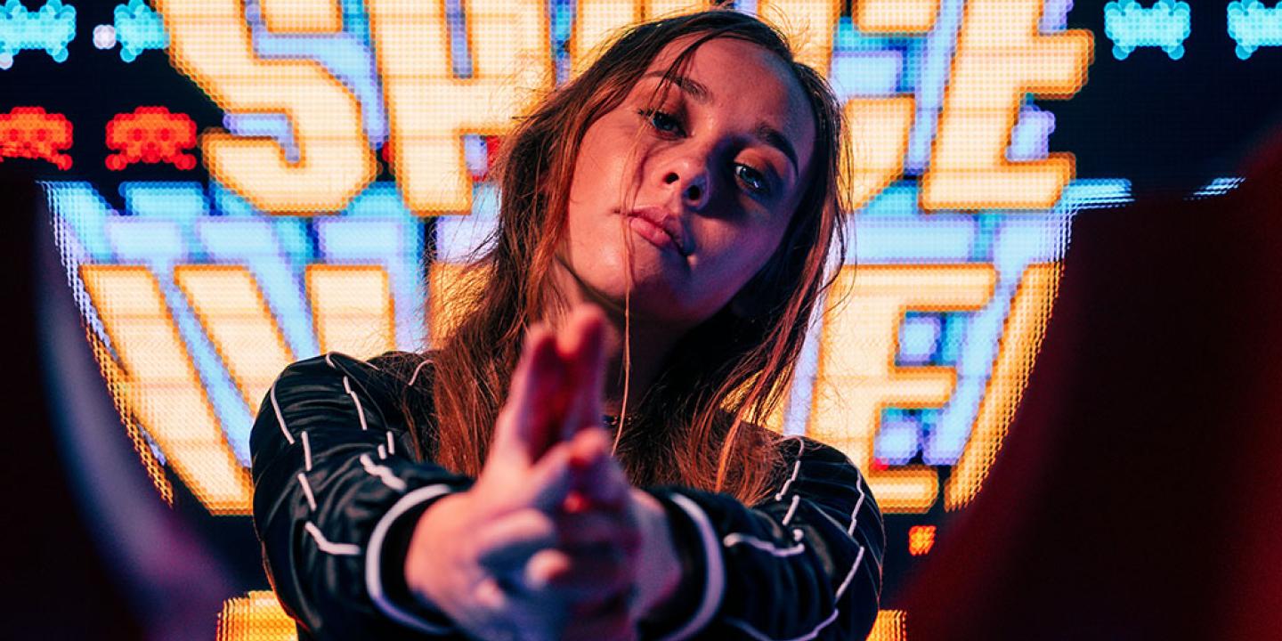 A woman stand in front of a large video screen displaying the Space Invaders title, hold her hands out in front of her.