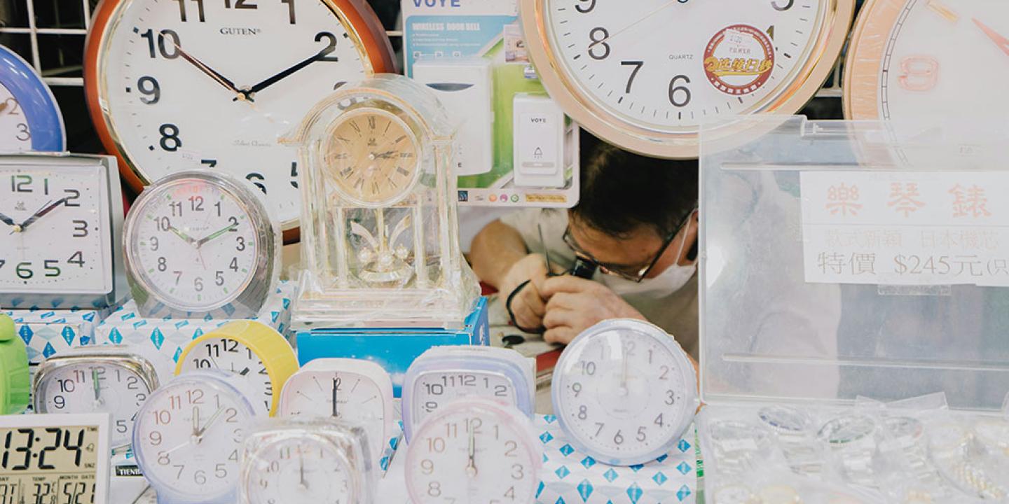 A clock repair peers at a clock he is repairing, amid a see of alarm and wall clocks on display