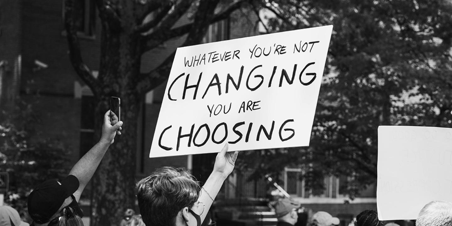 A protest placard is held above a march, reading 'If you don't change it, you choose it'.