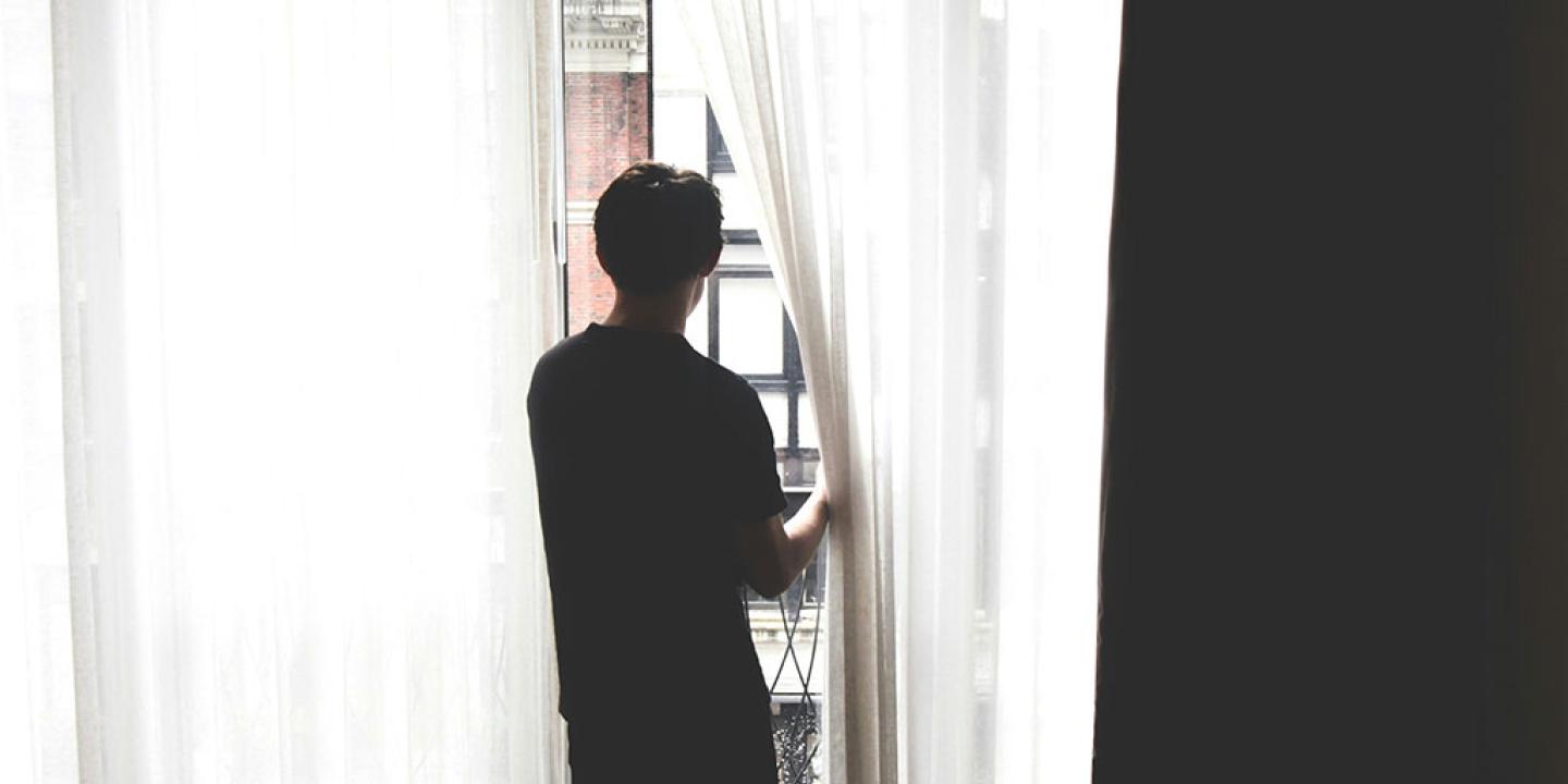 A persons stands, holding a net curtain aside to gaze out.